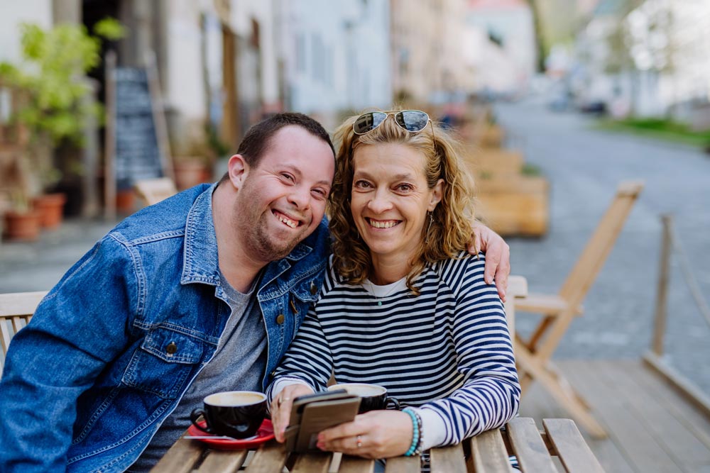 Portrait of happy young man with Down syndrome with his mother sitting at cafe outdoors and talking